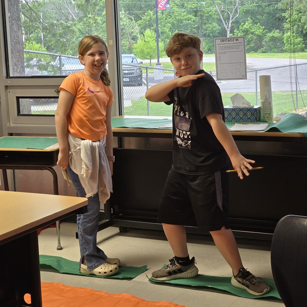 Two children, a girl and a boy, are standing on green mats in a classroom. A long desk is behind them.