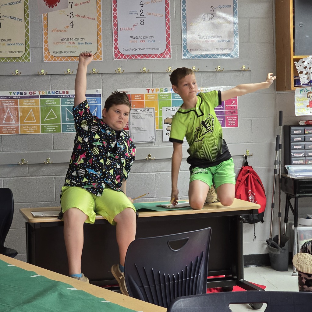 Two young boys standing on a table in a classroom. One boy is raising his arms.