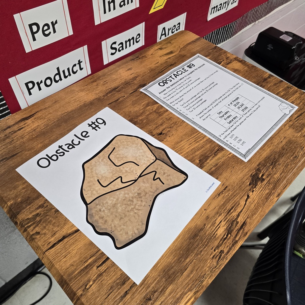 A wooden table displays a paper with a rock drawing and another paper with text. Behind the table is a red wall with text.