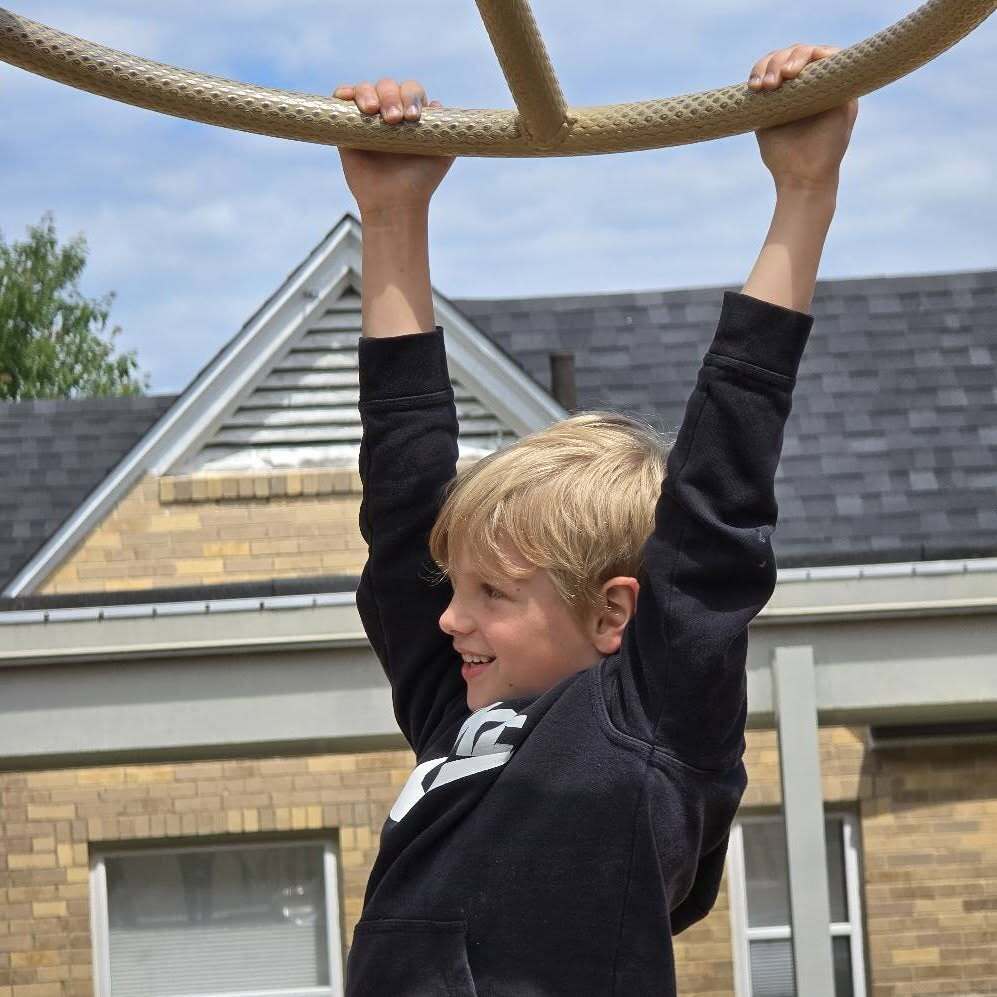 Child in black shirt hangs from a rope swing in front of a building with brick walls and gray roof.