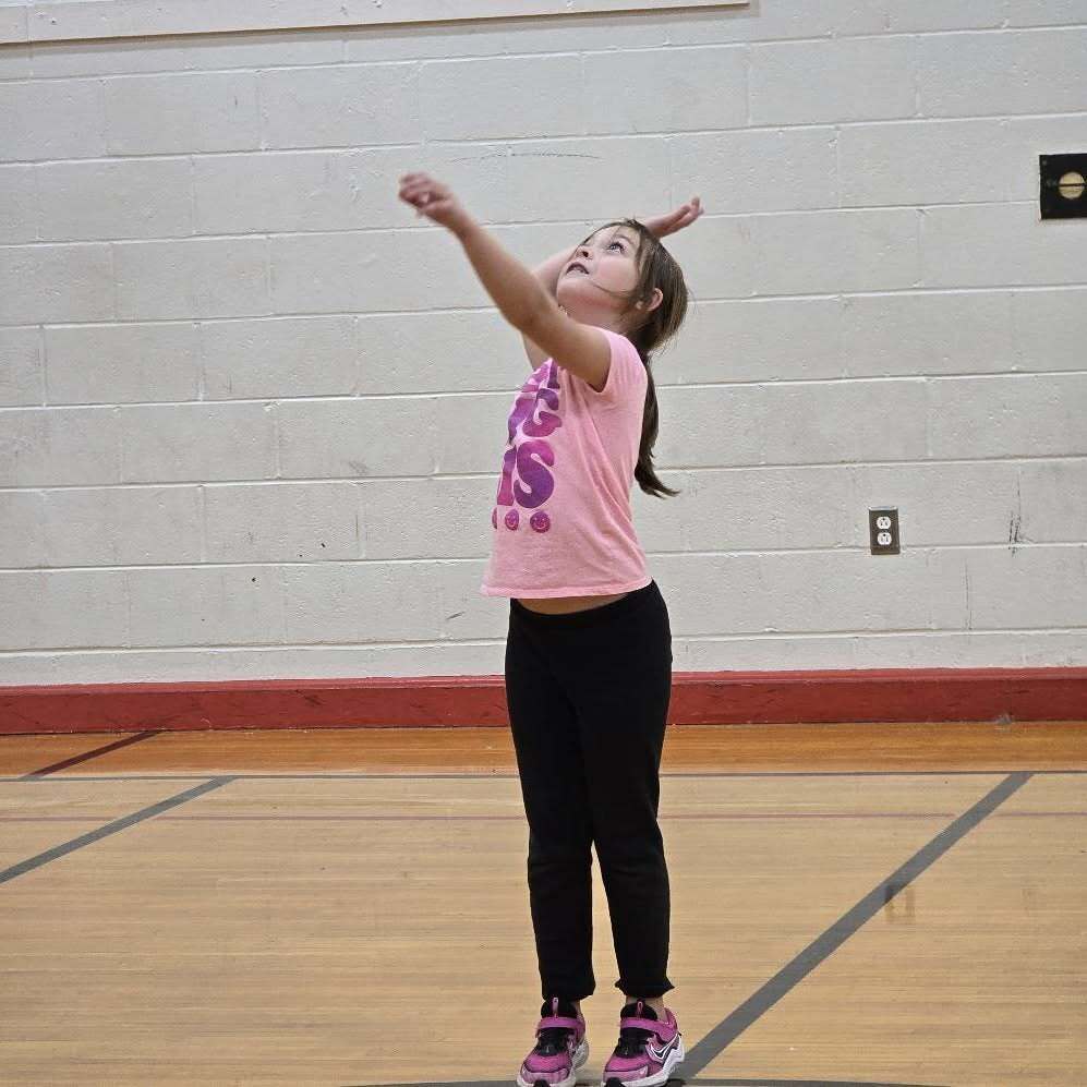 A young girl with pink shirt and black pants serves a ball in a gym.