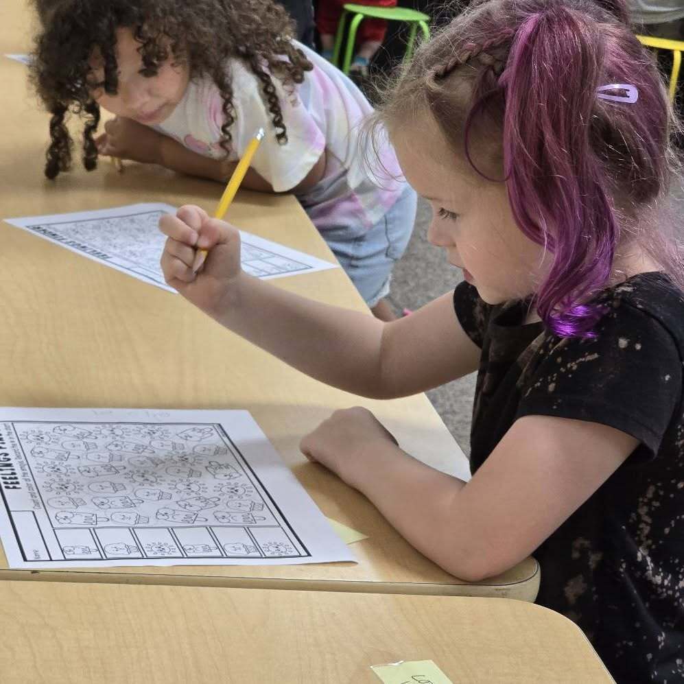 Two young girls in a classroom, one with purple hair and the other with curly hair, are both doing puzzles.