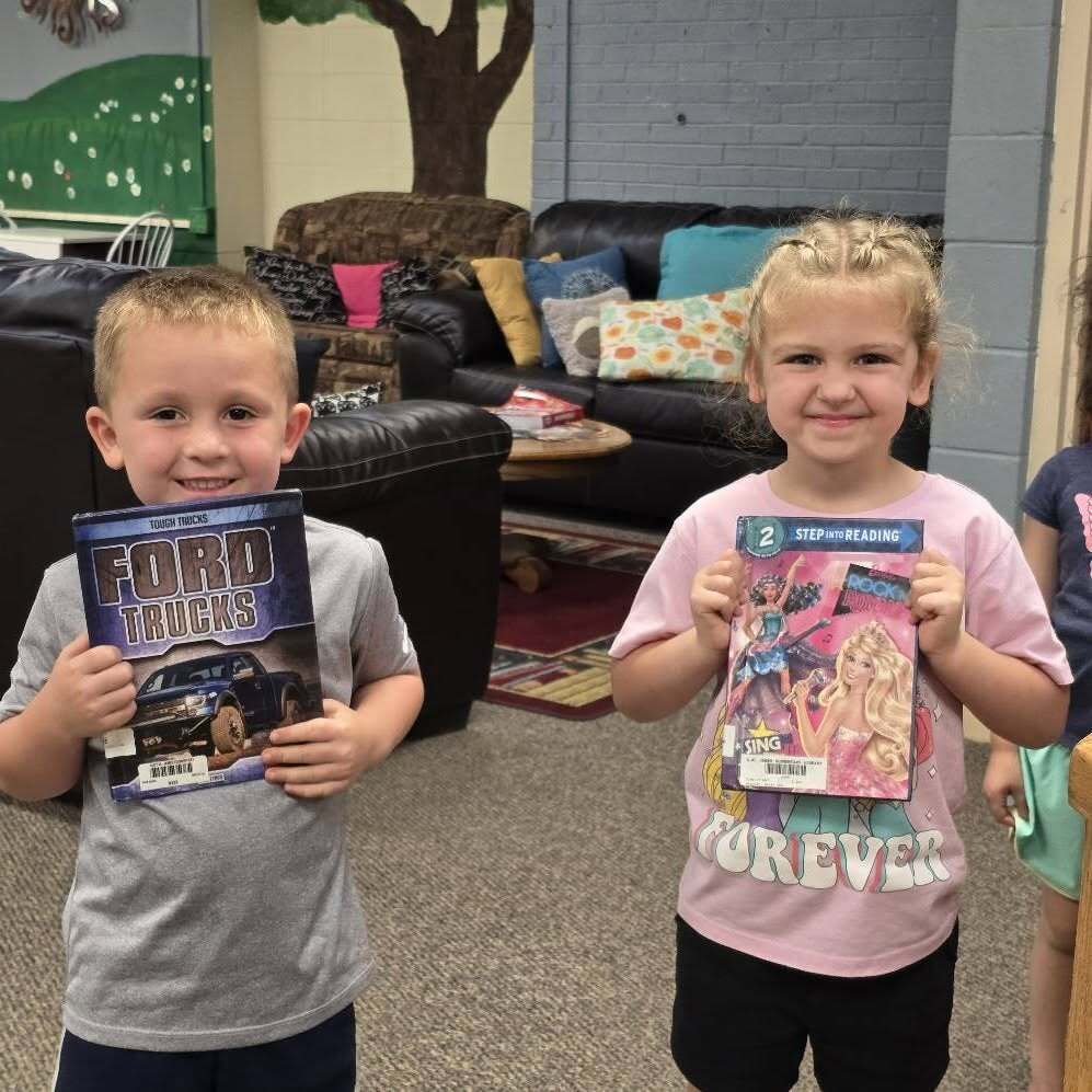 Two children stand next to each other, smiling. One holds a book titled "Ford Trucks." Behind them, a room has a green wall, a tree, and furniture.