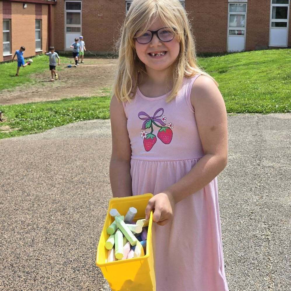 A girl with blonde hair holds a yellow container of chalk outside a school with a grassy area.