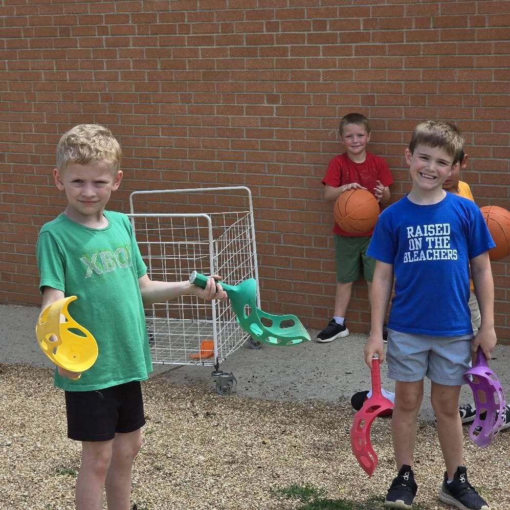 Three children hold toy helmets and balls in front of a brick wall and a small playground.
