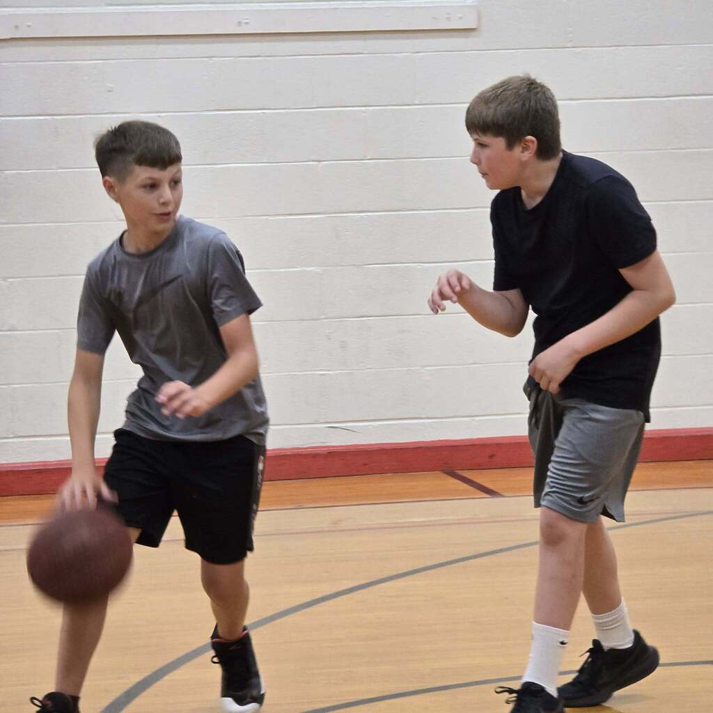 Two boys play basketball indoors; one dribbles, the other reaches out; court has red line.