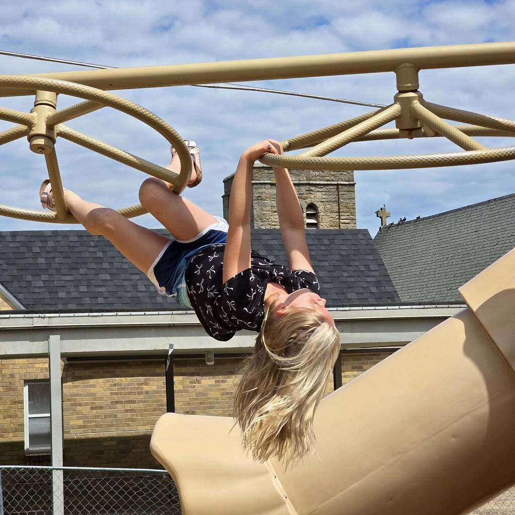 A person is upside down on a large climbing structure with a slide. Behind them is a building with a slate roof and a steeple.