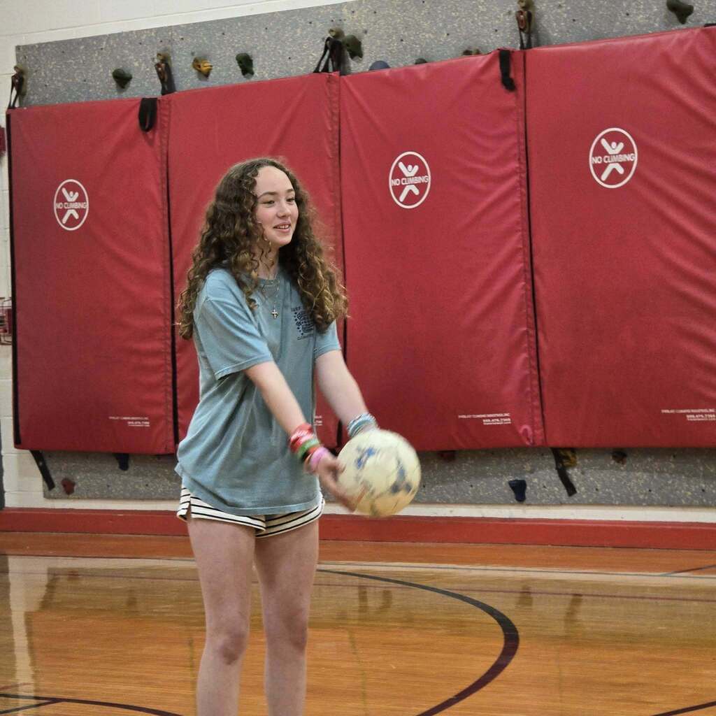 A young person practices holding a ball in an indoor gym. Red safety padding lines the wall behind.
