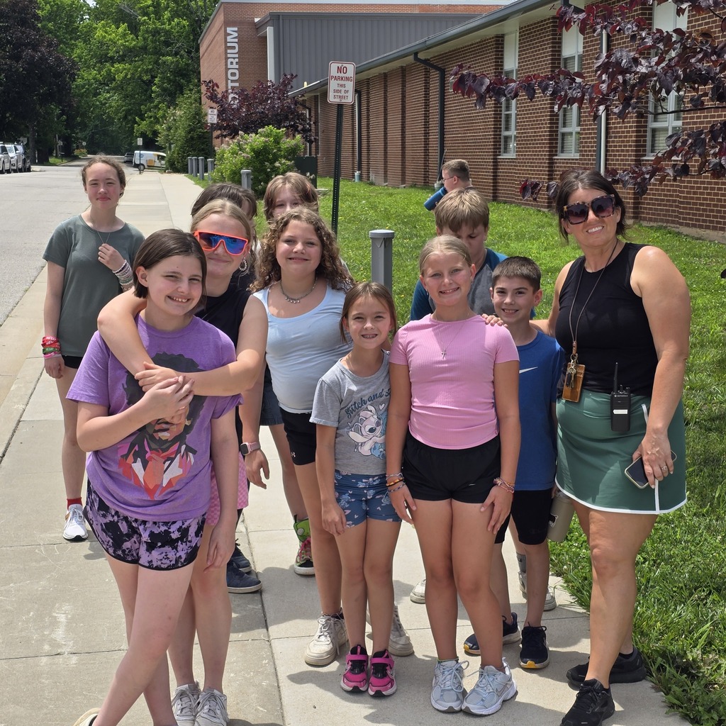 A group of children and an adult, wearing casual clothes, standing on a sidewalk in front of a building.