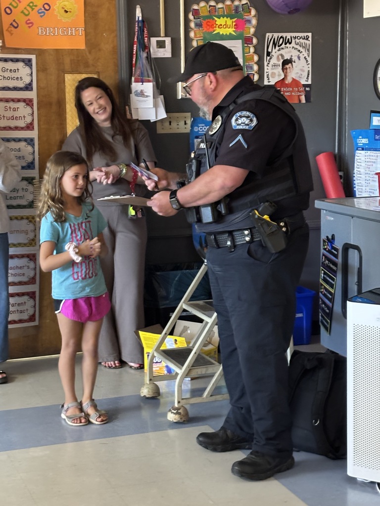 A police officer in uniform hands an item to a young girl with a bracelet, while a woman stands nearby. The scene is indoors with posters on the wall.