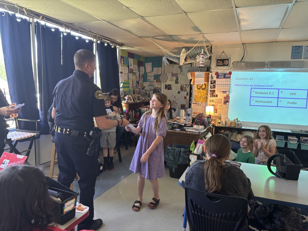 A police officer stands with a girl in a classroom, speaking to her. Other children sit at tables.