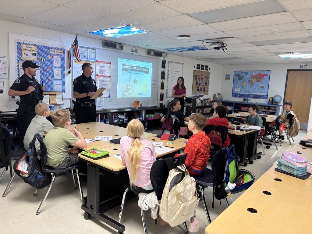Police officers stand by a projection screen in a classroom. Children sit at desks and listen.
