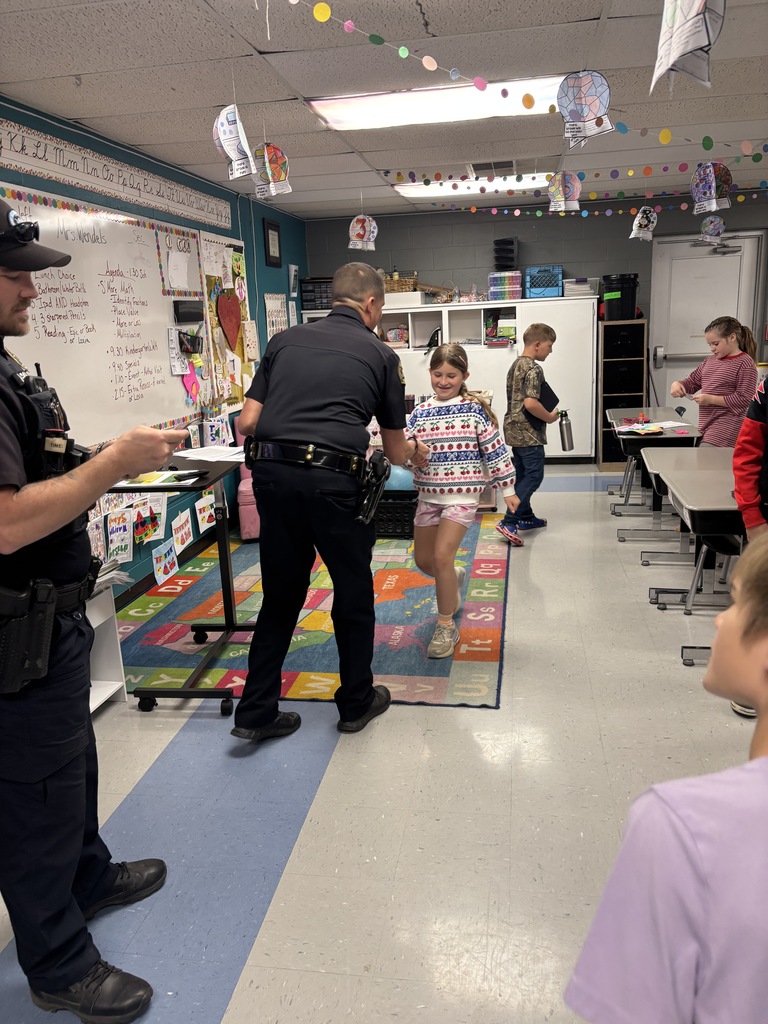 Two police officers talk with a child wearing a Christmas sweater in a classroom. Other children look on.