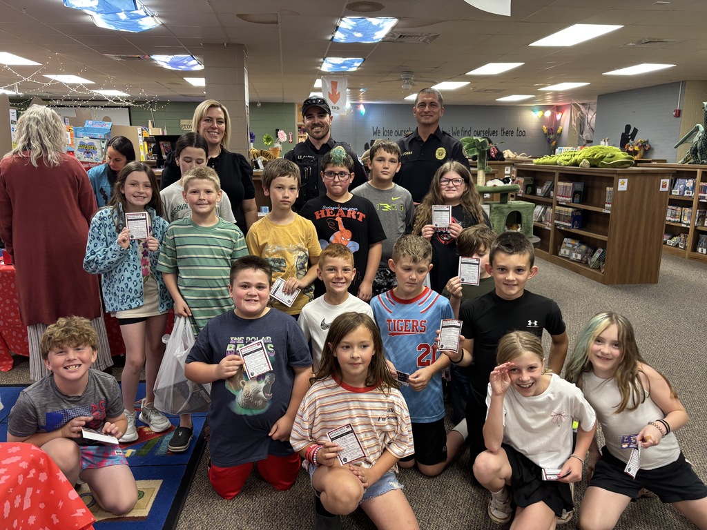 A group of children and adults in a room, likely a library. Some hold certificates, and some wear backpacks.