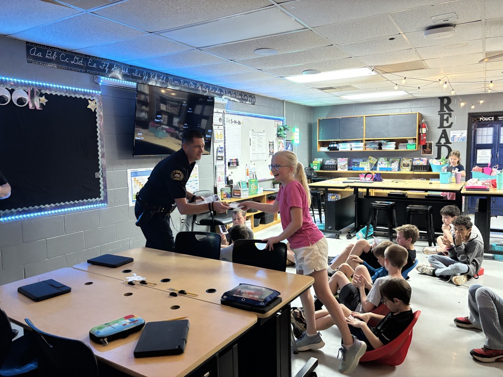 A police officer speaks to a girl and other students in a classroom, with tables and chairs.