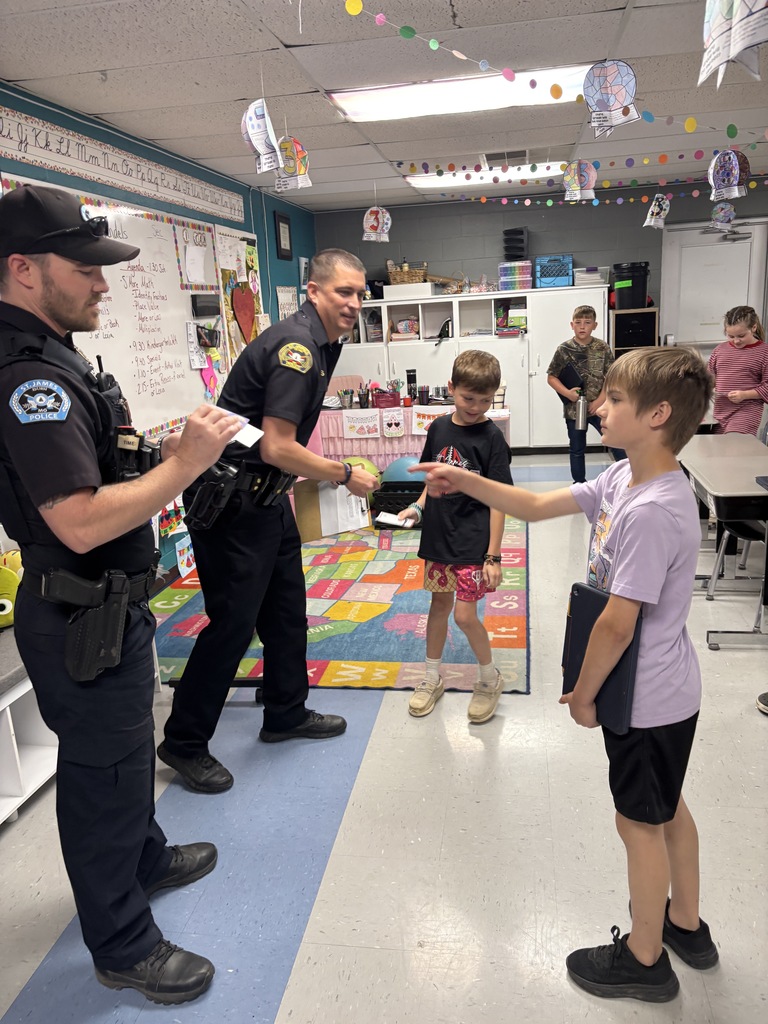 Two boys shake hands with police officers in a classroom with a colorful rug and whiteboards.