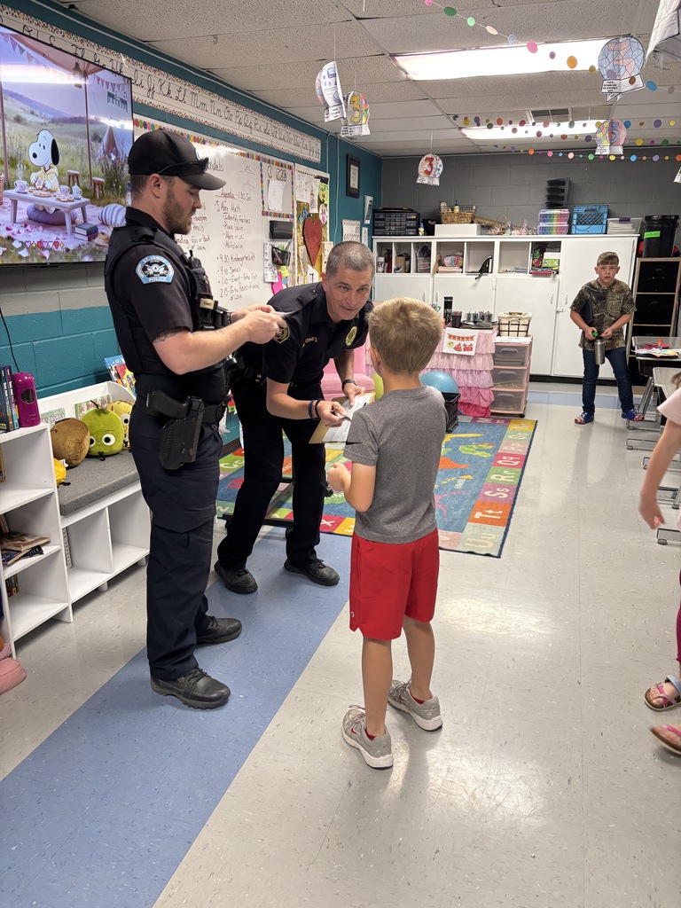 Police officers interact with a young boy in a classroom. Bright blue floor, white walls, and colorful decor.
