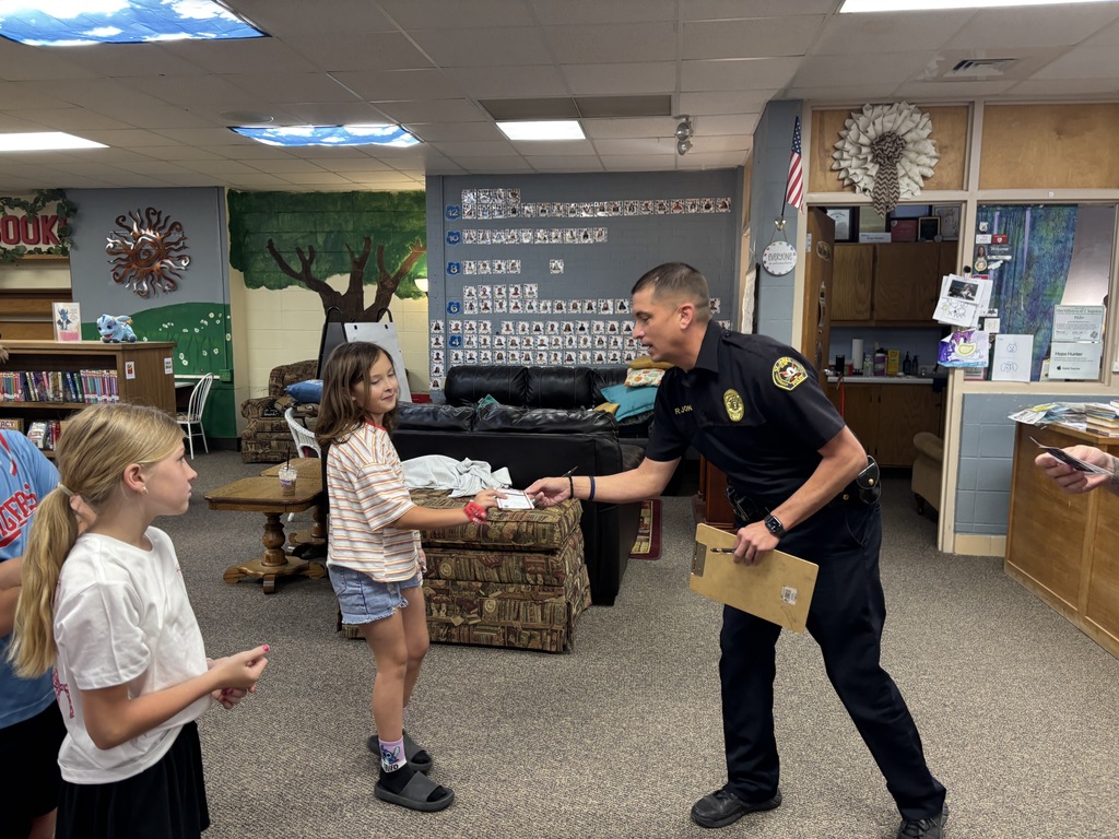 Police officer shakes hands with two young girls. One girl wears sneakers, the other sandals. Background includes shelves and a tree mural.