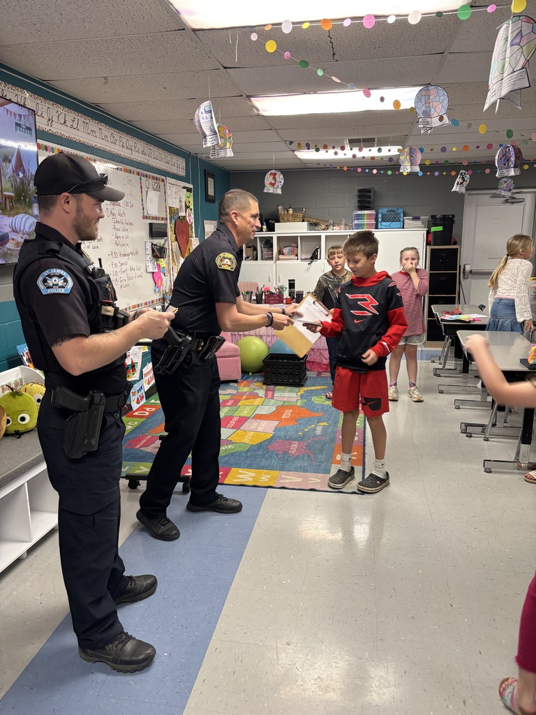 Two police officers interact with a young boy in a classroom setting; the boy holds a certificate.