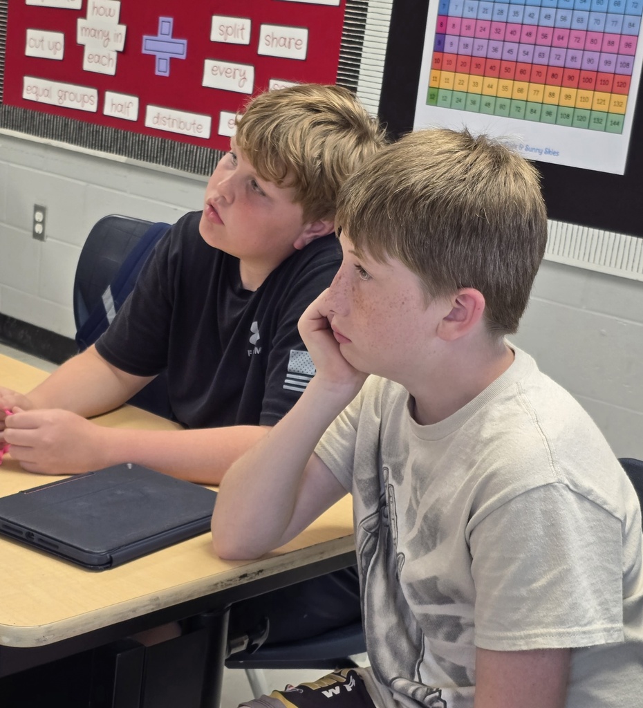 Two young boys seated at a desk in a classroom, one looking downward, another with hand on chin.