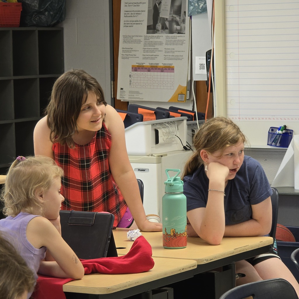 A classroom with several students. A woman with brown hair is standing next to a girl seated at a table.