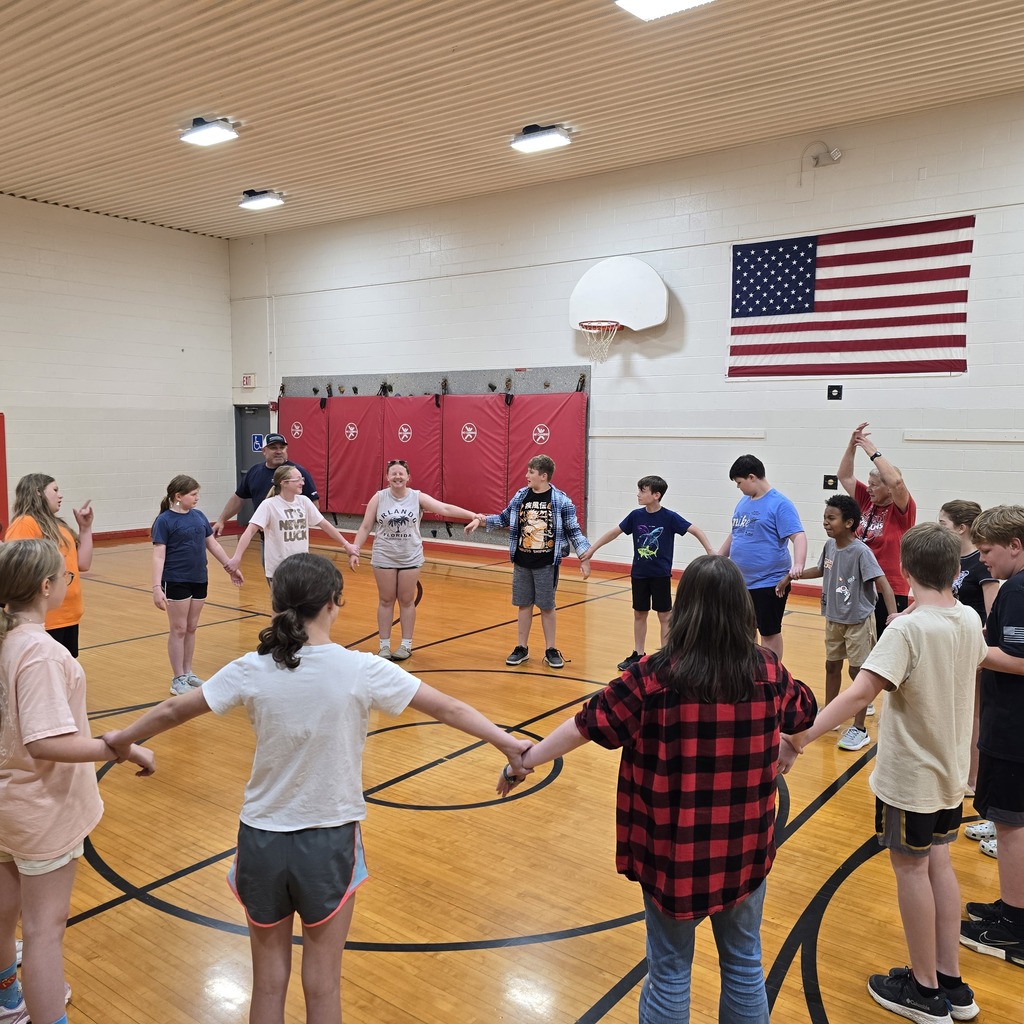 Children and adults are inside a gym, holding hands in a circle. A flag hangs on the wall.
