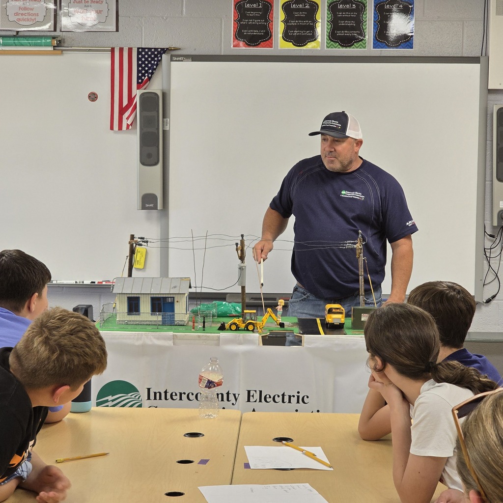 People sit at a table; a man in a blue shirt and cap stands in front of a model.