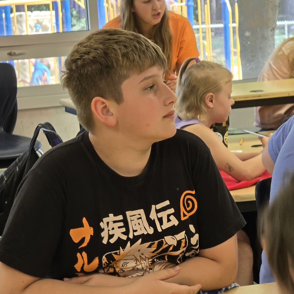 A boy wearing a black T-shirt sits with arms crossed. Behind him, a girl writes. They are near a window showing a playground.