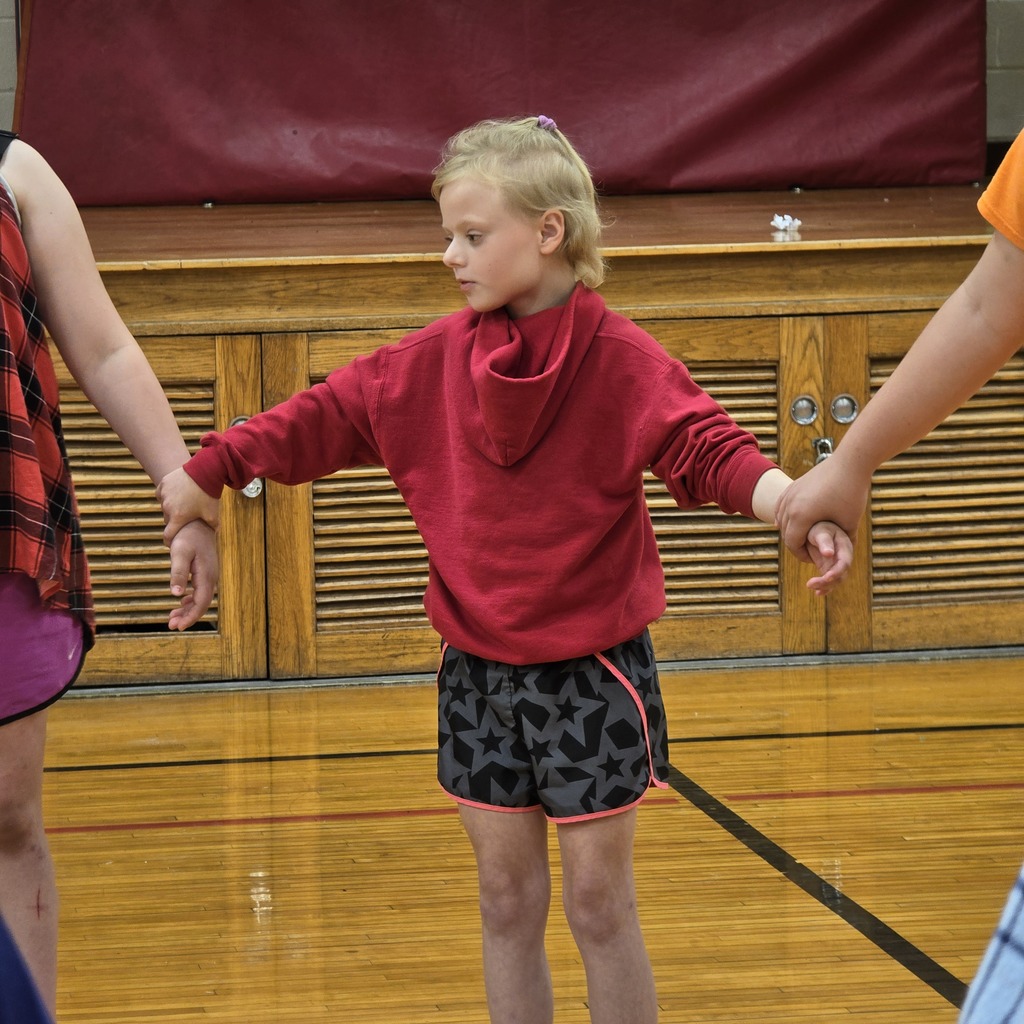 A young girl with short blonde hair holding hands with two people in a gymnasium.