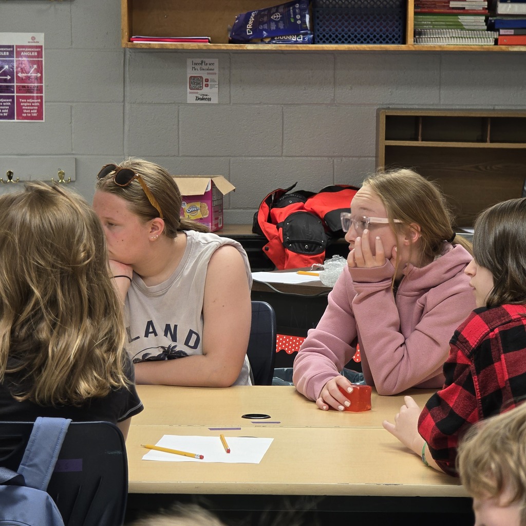 Five students in a classroom, one with glasses and a pink sweater, another in a white shirt.