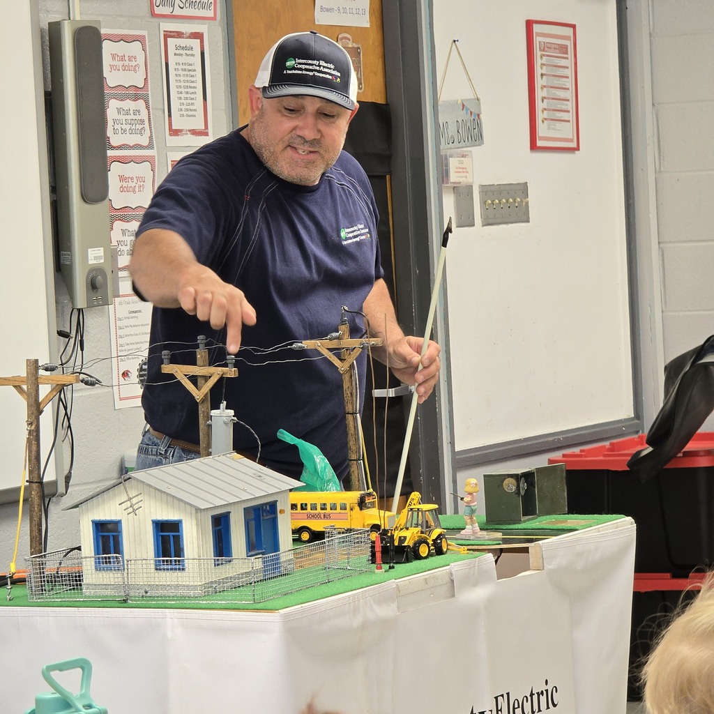 Man in a blue shirt pointing at a miniature model of a house and utility poles.