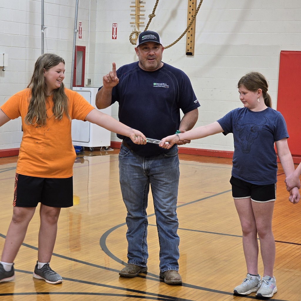 Three people in a gym hold a stick in their hands. A man gestures with his index finger.