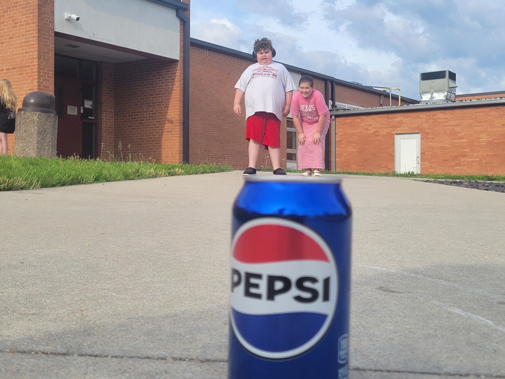 Two children stand behind a can of Pepsi, with a brick building in the background.