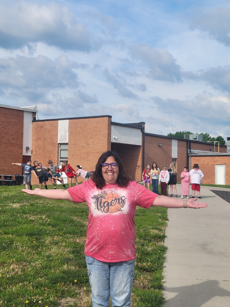 Woman in pink shirt stands with arms wide open in front of building. Children in red shirts stand in background.