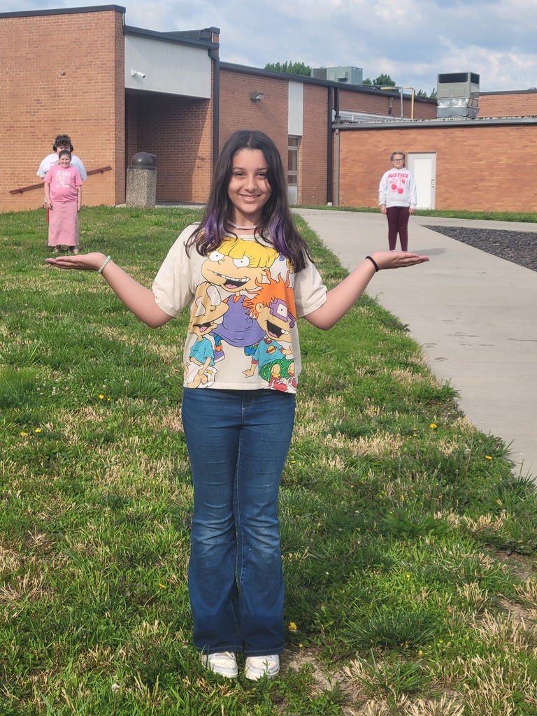 A person stands with arms outstretched on a grassy field, wearing a colorful t-shirt and jeans.