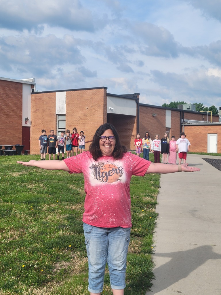 A woman in a pink shirt stands on a sidewalk with arms wide open, near a school building.