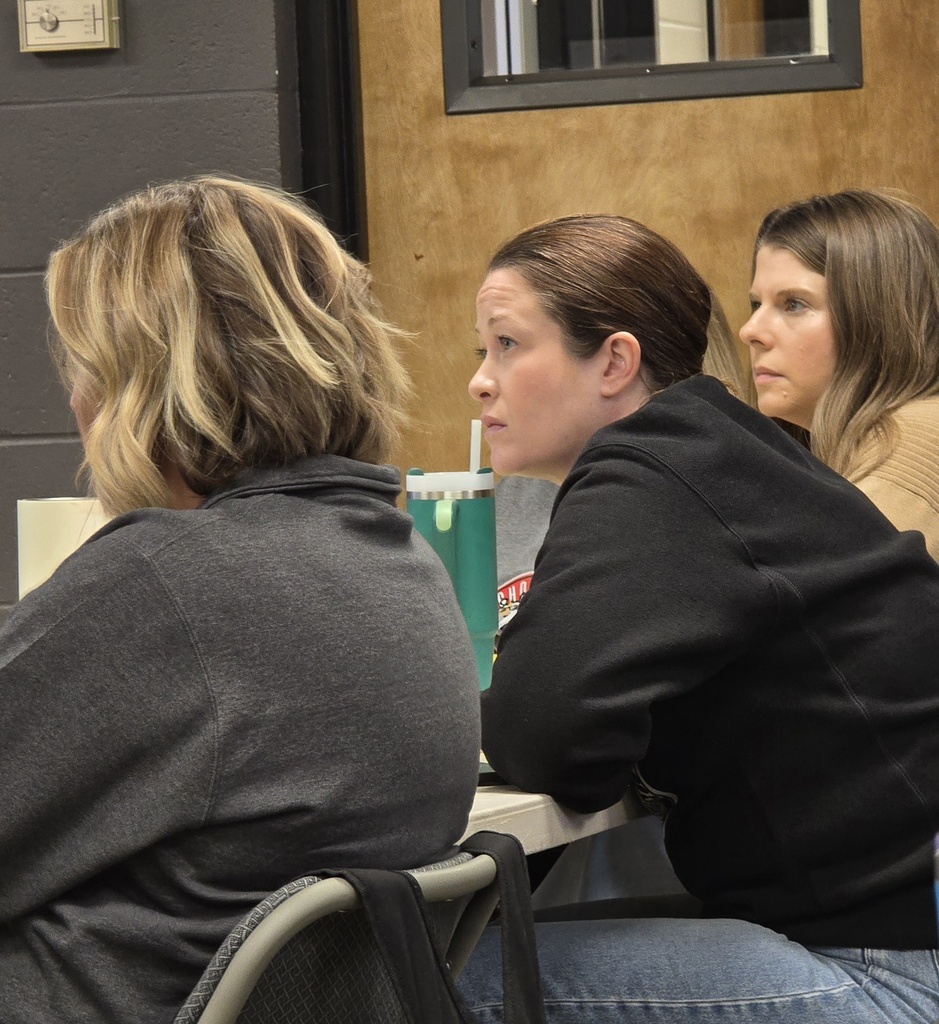 Three women seated in a classroom with a cup and straw on a table between them.