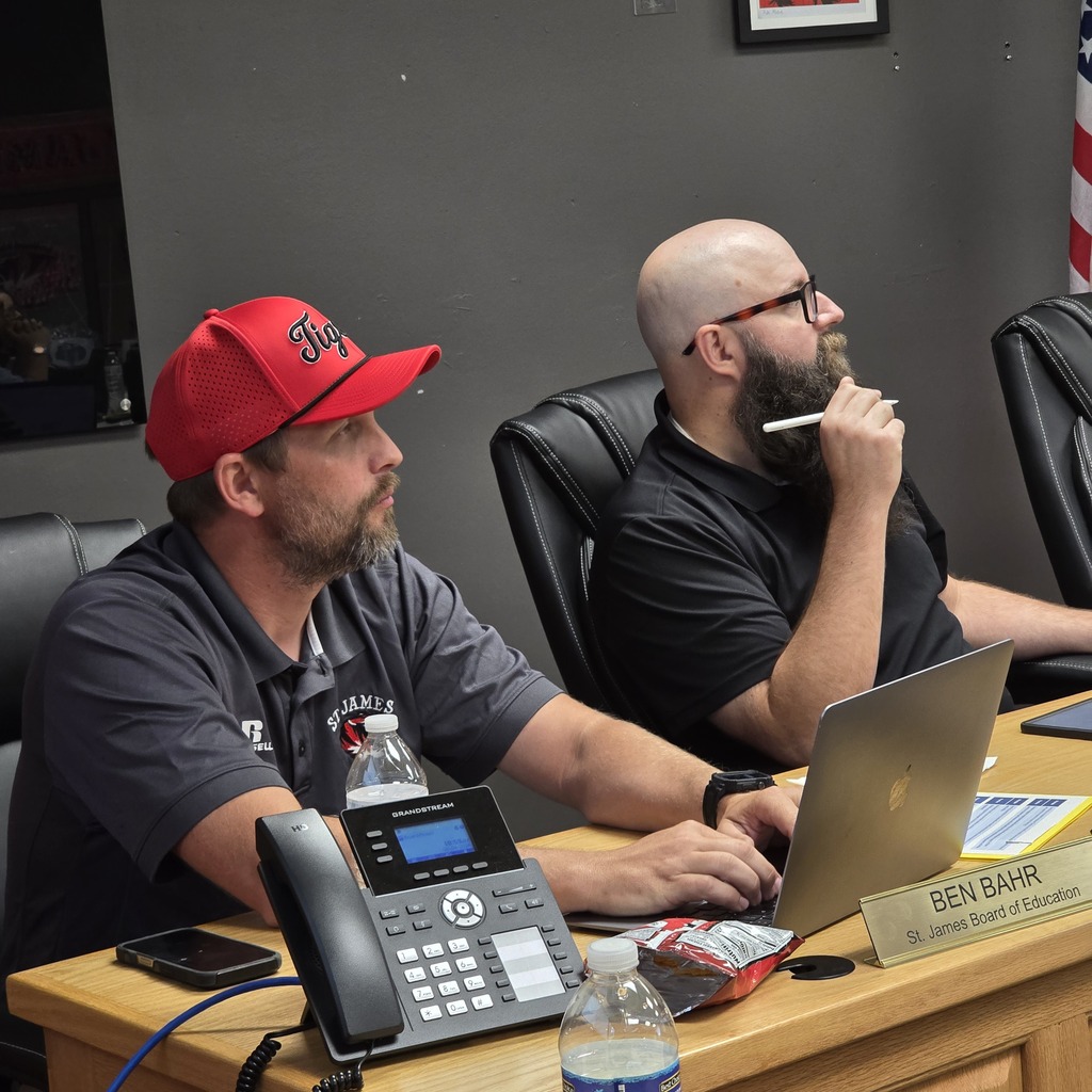 Two men sit in a room. One in a red hat looks at a laptop. The other wears glasses and smokes.