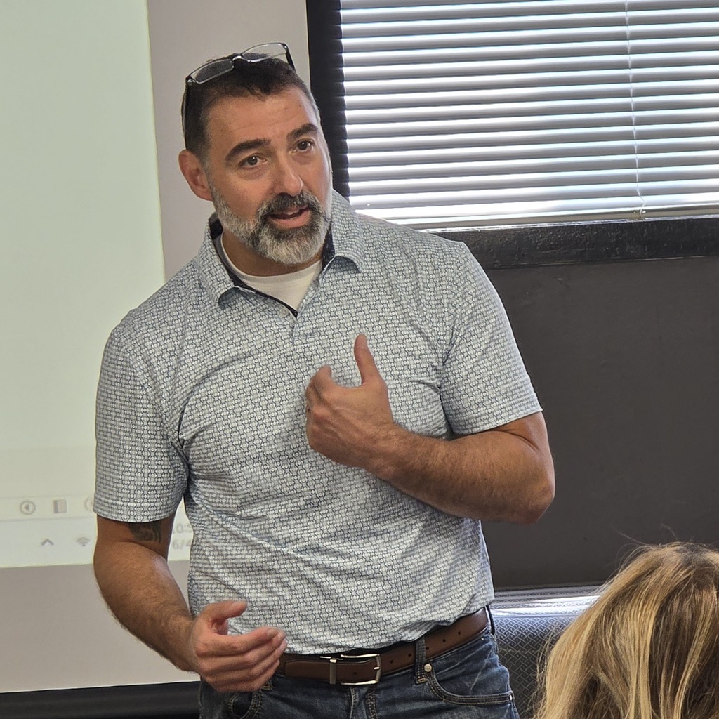 A man speaks in a classroom. He wears a grey shirt and jeans. He gestures with one hand.