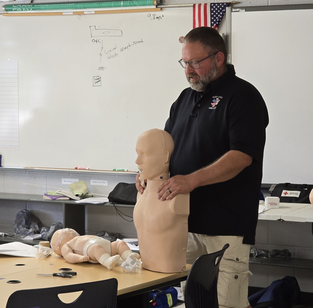 A man in a classroom demonstrating CPR techniques on a dummy, with an infant dummy and a whiteboard in the background.