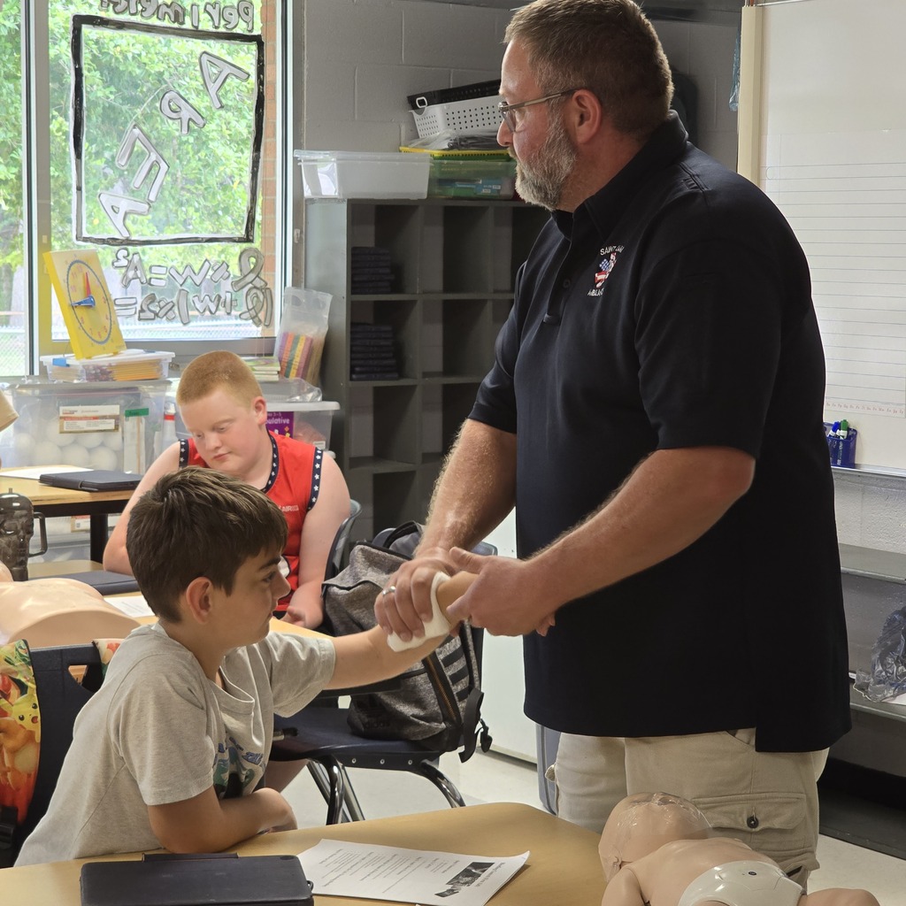 A man stands, demonstrating a skill to a boy seated at a table in a classroom.