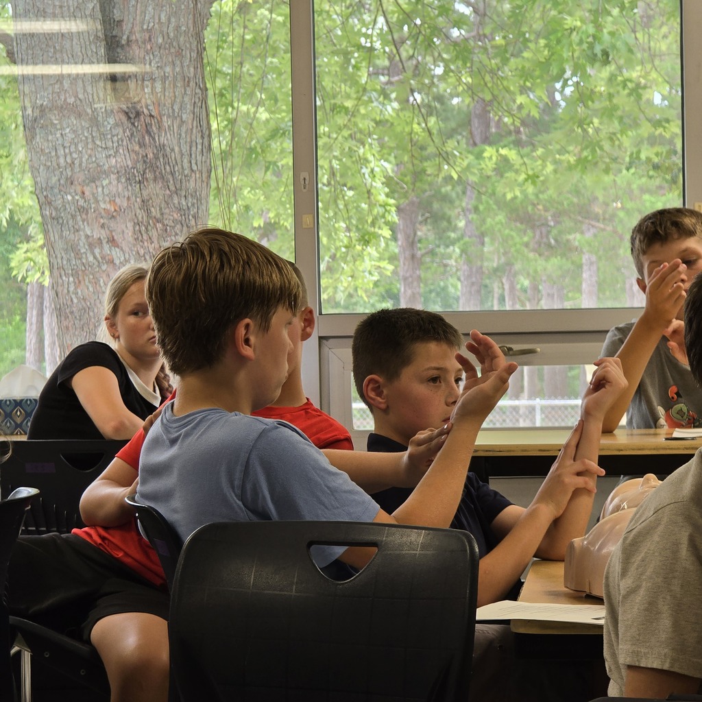 Five children, seated and standing, engage in animated gestures and conversation in a classroom with large windows.