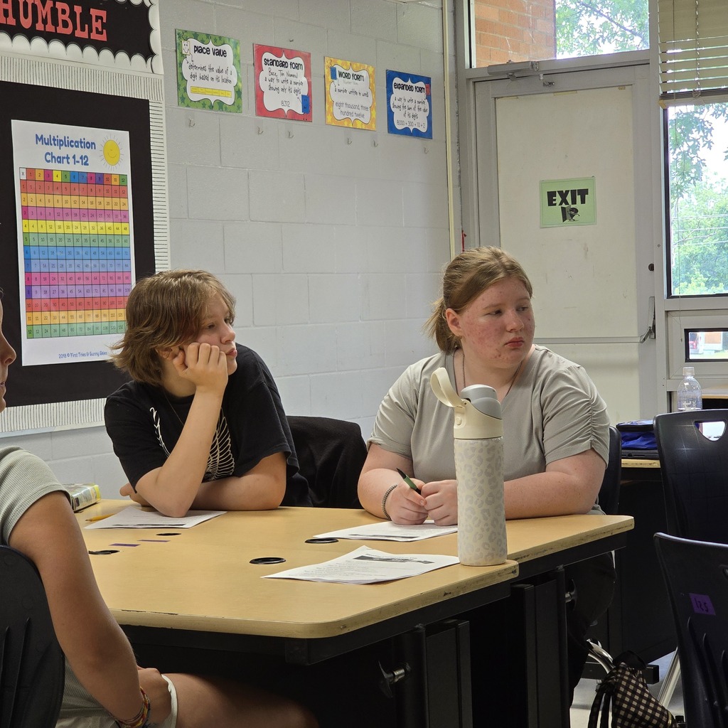 Two women sit at a table inside a classroom, one holding a pen. A sign says "Humble" and an exit sign.