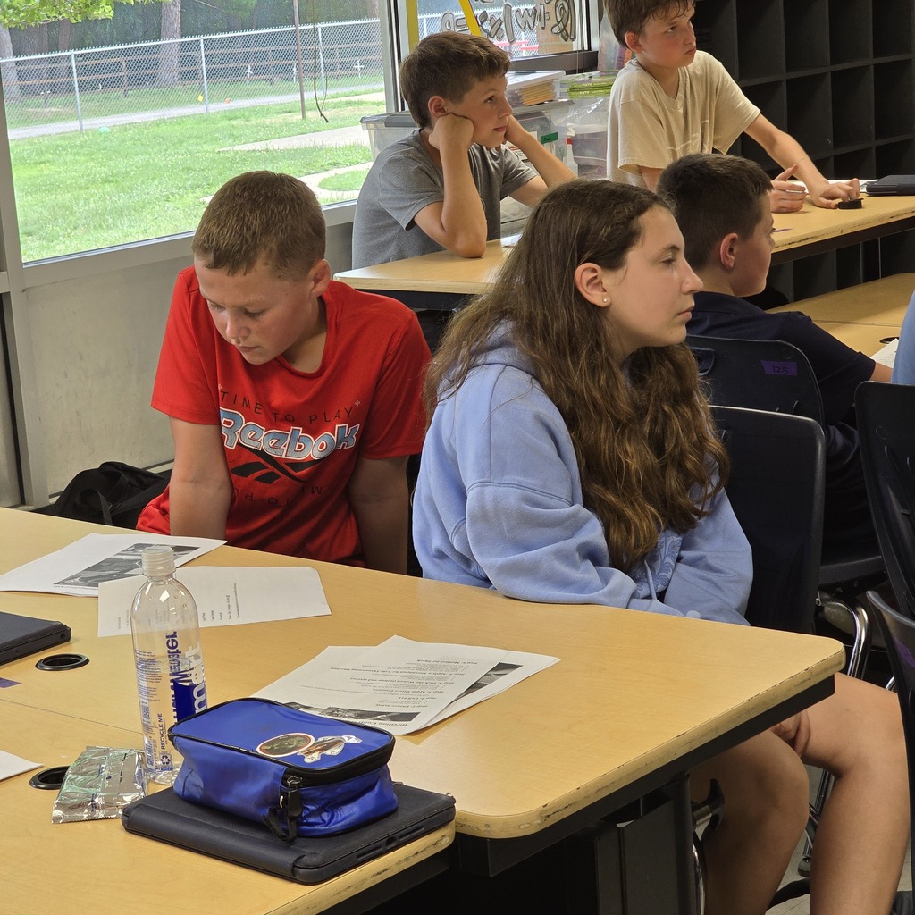 Several students sit at a table inside a classroom with a window showing a grassy area.