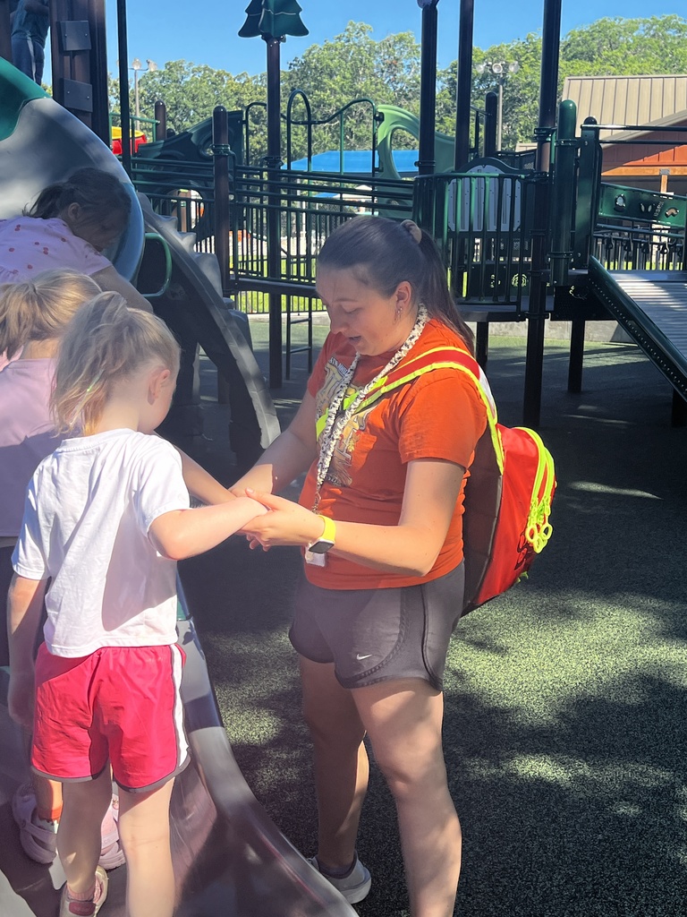 A young child stands with an adult at a playground. The adult holds the child's hand, and the child looks down.