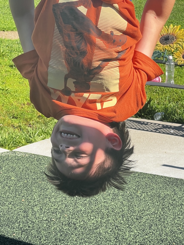 A child wearing an orange t-shirt is upside down on a playground surface with green grass and a yellow flower in the background.