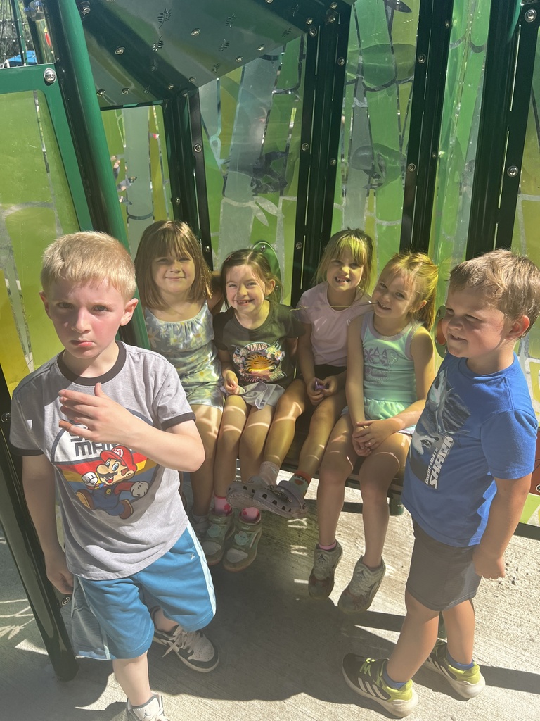 Children gather in a playhouse. Four sit on a bench; two stand. Green background with reflections of trees.