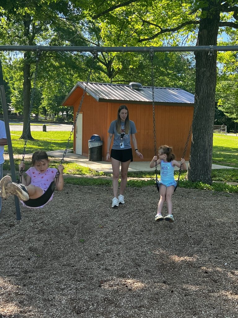 Two children are on swings at a park. A woman stands between them, holding a swing. A building is in the background.