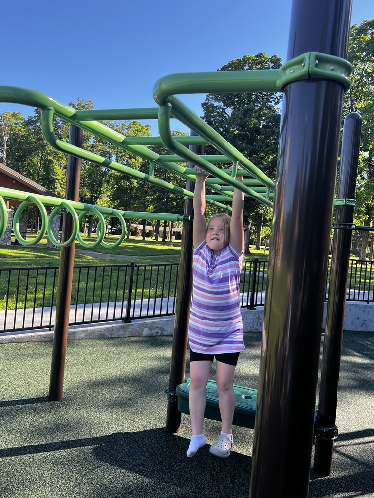 A young child stands on a pull-up bar in an outdoor playground. Green rings are above and the ground is rubbery.
