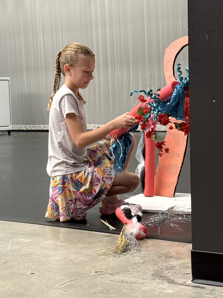 A young girl kneels on a stage, using her hands to adjust a colorful arrangement of fabric and objects.